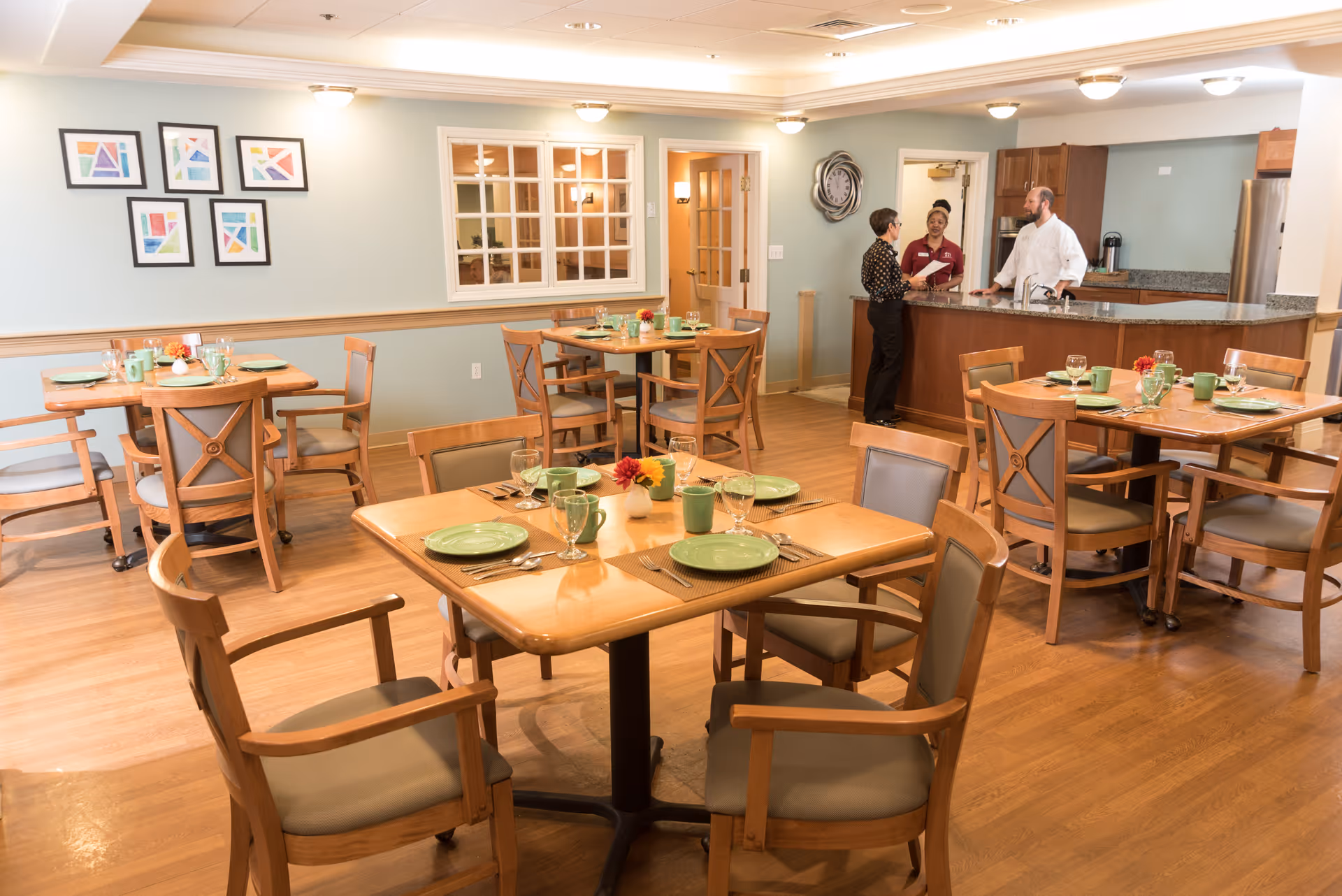 A dining room in a senior living facility with wooden tables and chairs set with green plates, cups, and glassware. Three staff members are standing and talking near a kitchen counter in the background. The room has light blue walls, framed colorful artwork, and a wall clock.