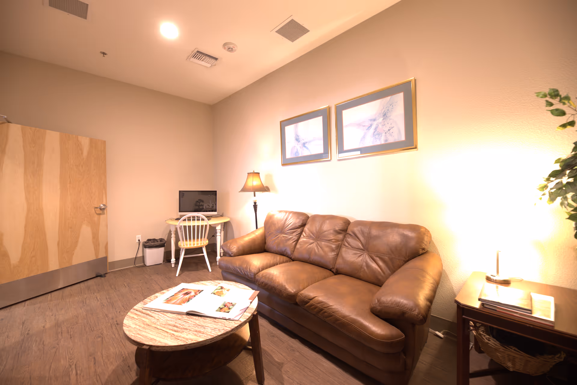 A cozy room with a brown leather couch, a wooden coffee table with an open magazine, a small desk with a computer and chair, a floor lamp, and two framed pictures on the wall. The room has wooden flooring and beige walls.