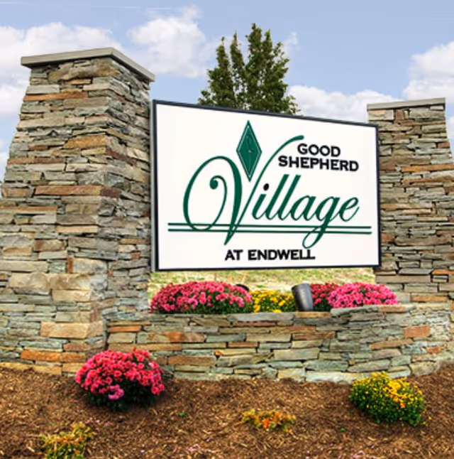 Stone monument sign for Good Shepherd Village at Endwell surrounded by flower beds with pink and yellow flowers, set against a backdrop of a tree and blue sky with clouds.
