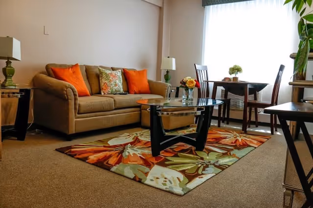 A cozy living room with a beige sofa adorned with orange and floral cushions, a glass coffee table on a colorful floral rug, two green table lamps on side tables, and a small dining table with two chairs near a large window with sheer curtains.
