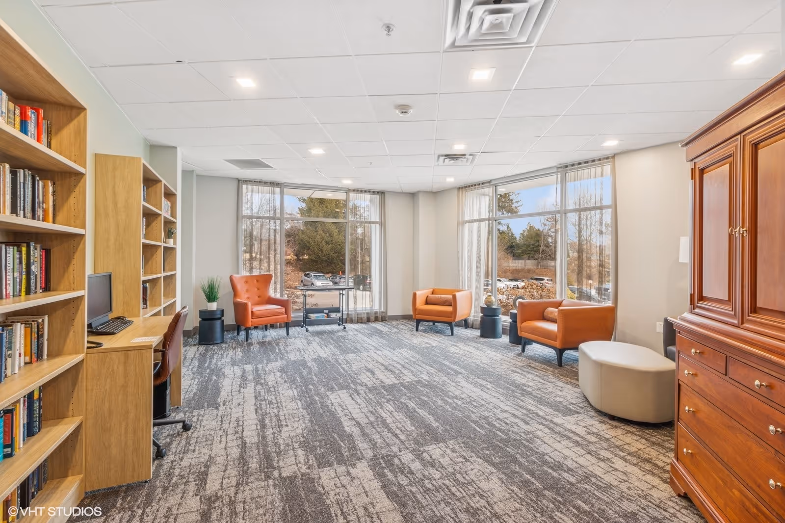 A bright and spacious room with large windows letting in natural light. The room features a carpeted floor, three orange armchairs, a wooden armoire, a small round ottoman, and built-in wooden bookshelves with a computer workstation. Outside the windows, trees and parked cars are visible.