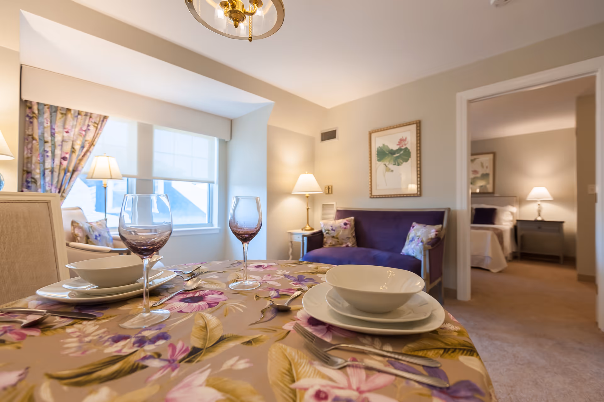A cozy interior of an assisted living facility showing a dining table set with plates, bowls, cutlery, and two wine glasses on a floral tablecloth. In the background, there is a purple loveseat with floral cushions, a side table with a lamp, and a framed botanical print on the wall. Through an open doorway, a bedroom with a bed, nightstand, and lamp is visible.