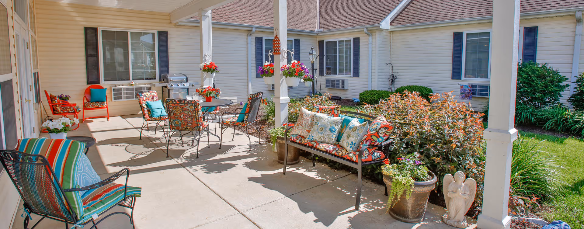 Outdoor patio area at Bickford of Lafayette with several colorful cushioned chairs and a bench around metal tables. There are potted plants and flowers hanging from the white pergola, with a barbecue grill near the building. The patio is adjacent to a garden with bushes and grass, and the building has beige siding with windows and air conditioning units.