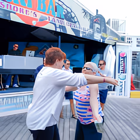 Two people outdoors, one with short red hair wearing a white shirt and the other with long gray hair wearing sunglasses and a striped sleeveless top, appear to be dancing or interacting playfully on a wooden deck with a colorful mural in the background and other people standing nearby.