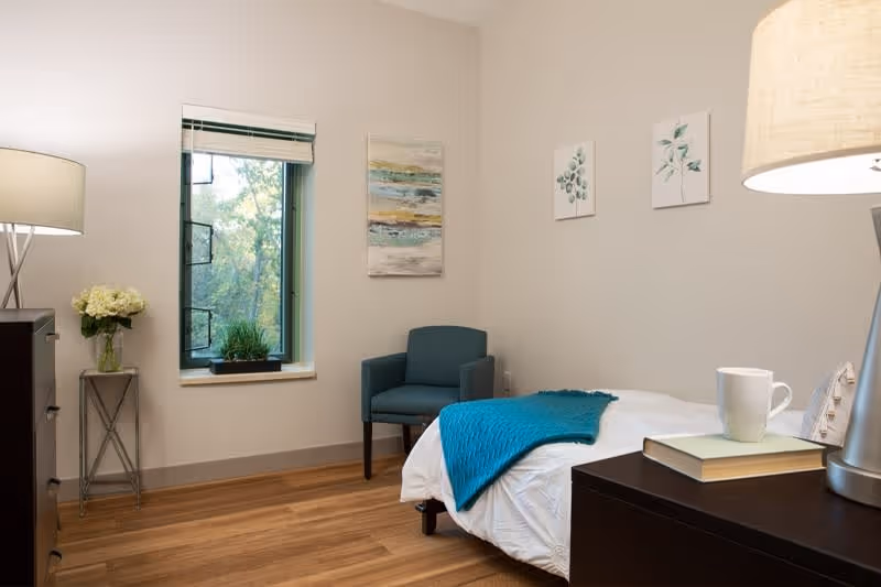 A cozy bedroom with a single bed covered in white bedding and a teal throw blanket. There is a dark wooden nightstand with a lamp, a book, and a white mug on it. A teal armchair is placed near a window with a view of green trees outside. The walls are decorated with three pieces of artwork featuring abstract and botanical designs. A small table with a vase of white flowers is next to the window.