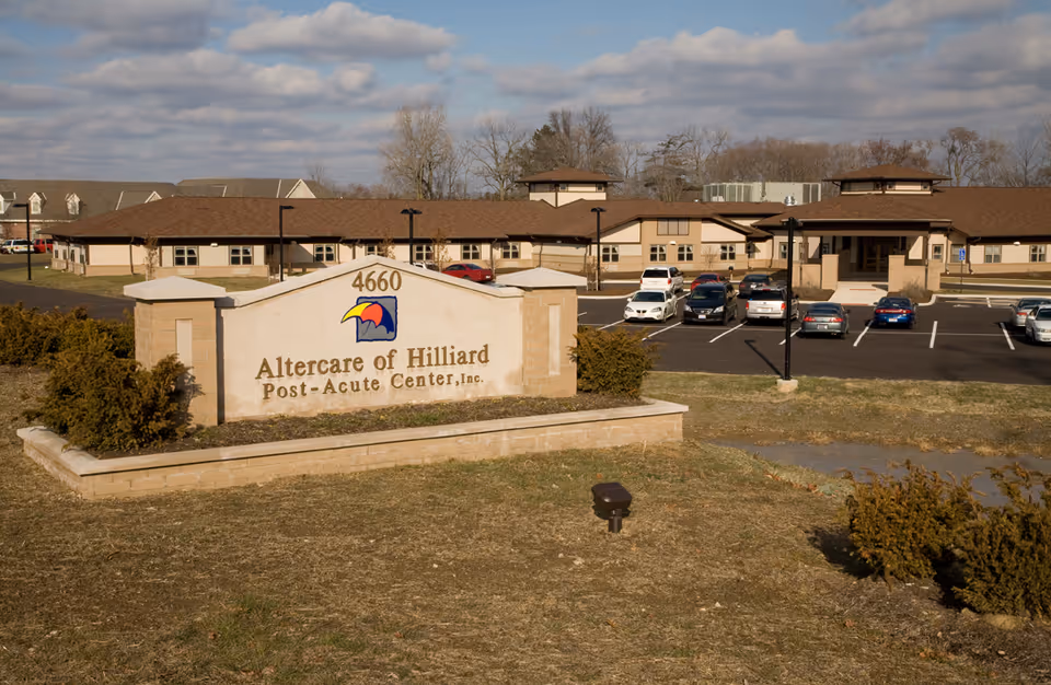 Exterior view of Altercare of Hilliard Post-Acute Center building with a parking lot and several parked cars in front. A large sign with the facility name and address 4660 is visible in the foreground, along with some bushes and grass under a partly cloudy sky.