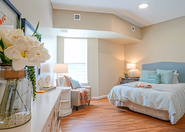 A bright and cozy bedroom with a large bed featuring light blue and white bedding, a bedside table with a lamp, a comfortable armchair with a throw blanket, and a window with blinds allowing natural light to fill the room. A vase with white flowers is visible on a dresser in the foreground.