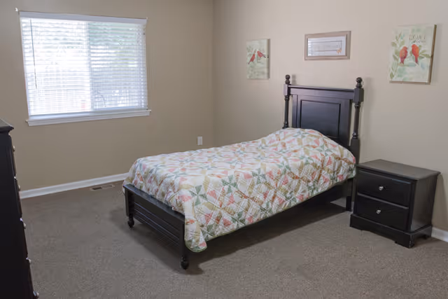 A simple bedroom with a single bed covered in a patterned quilt, a black wooden headboard, a matching black nightstand with two drawers, a window with white blinds, and three framed pictures on the beige walls.