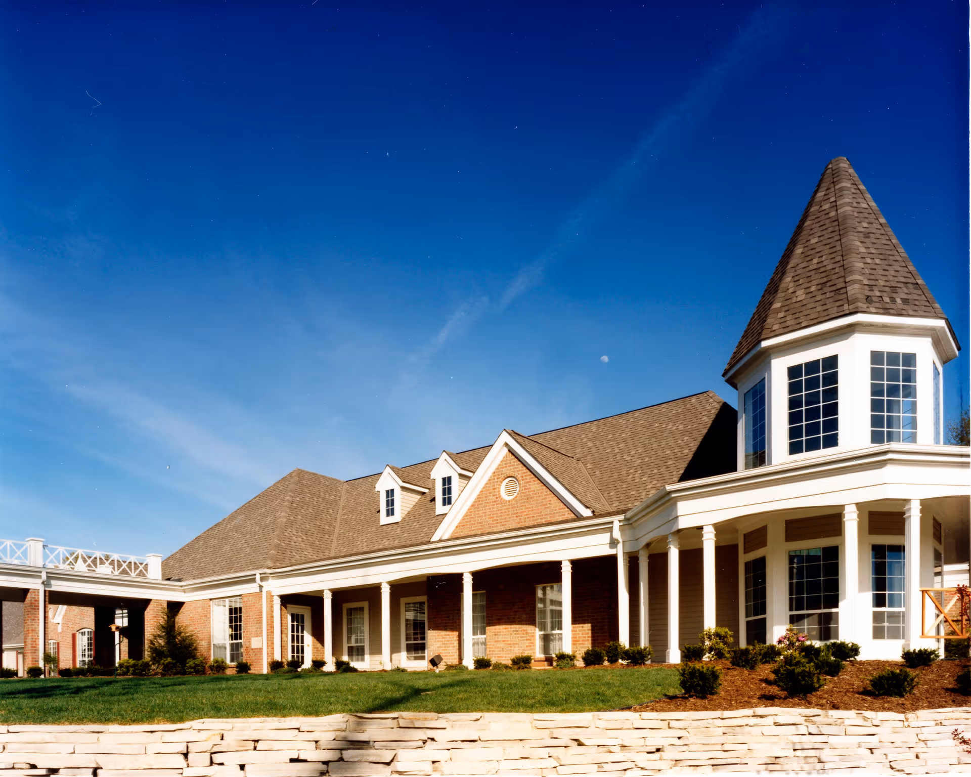 Exterior view of a senior living facility building with a brick facade, white columns, and a pointed turret under a clear blue sky.