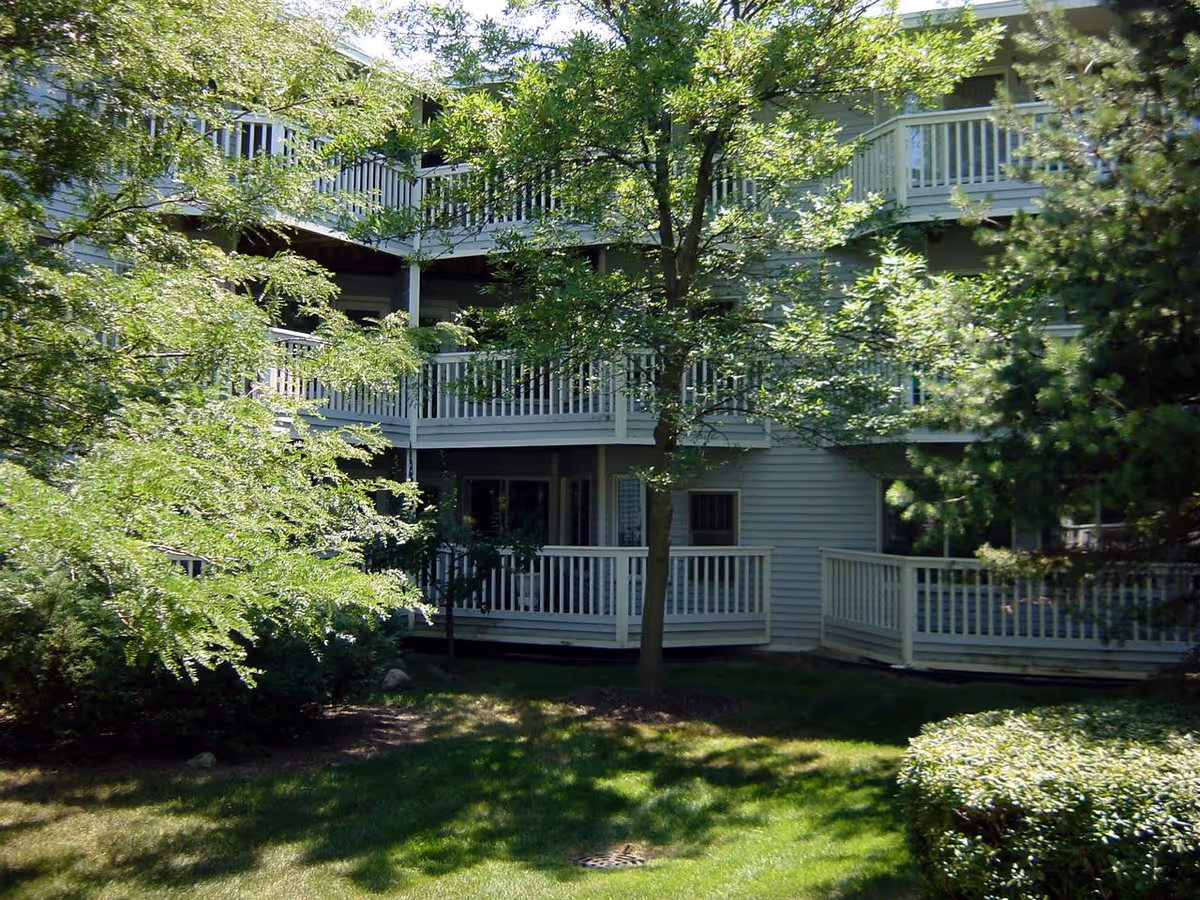 Exterior view of a senior living facility with multiple balconies surrounded by lush green trees and well-maintained grass.