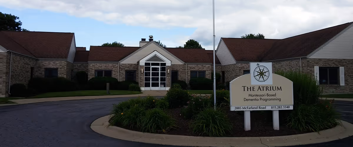 Exterior front view of The Atrium, a single-story brick building with a peaked roof and a central entrance with glass doors. In front of the building is a landscaped roundabout with green plants and a sign that reads 'The Atrium Montessori-Based Dementia Programming 2885 McFarland Road 815.282.3340'. The sky is partly cloudy.