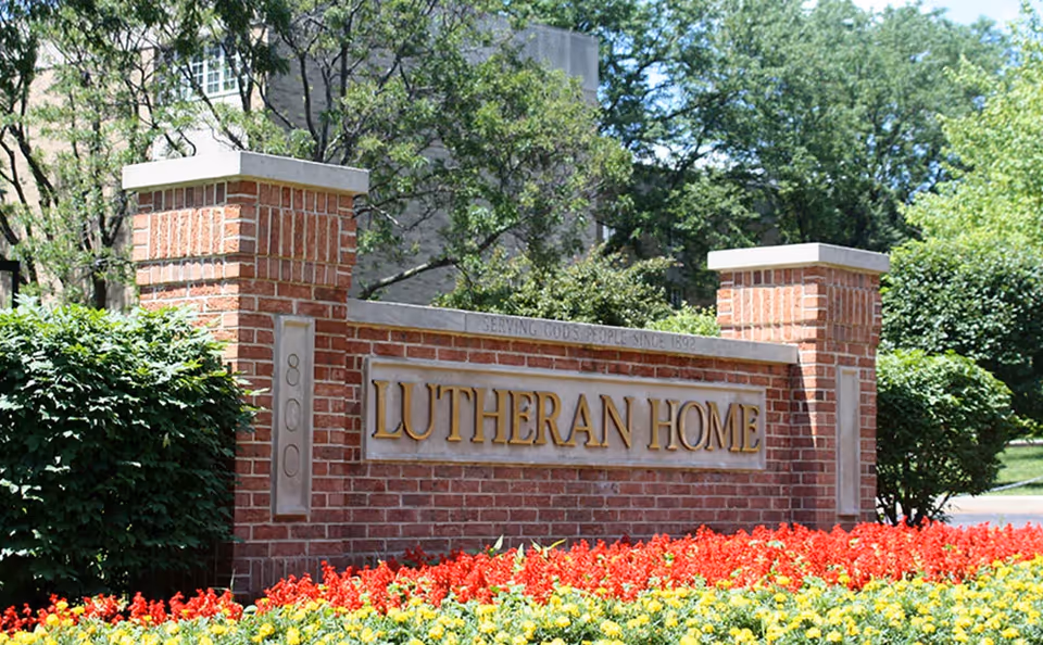 Brick sign for Lutheran Home surrounded by green bushes and colorful red and yellow flowers, with trees and a building in the background.