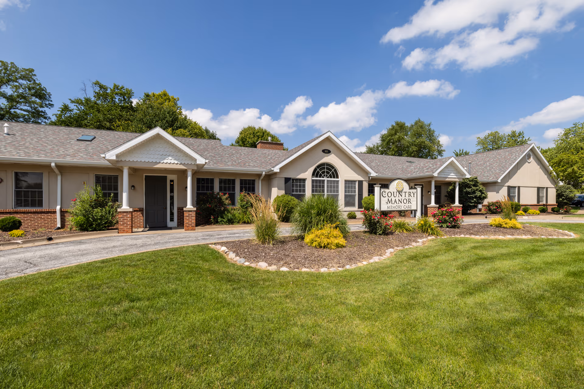 Exterior view of Country Manor Memory Care facility showing a single-story building with beige walls, a gray roof, and multiple windows. The building is surrounded by well-maintained landscaping with green grass, bushes, and flowers under a blue sky with some clouds.