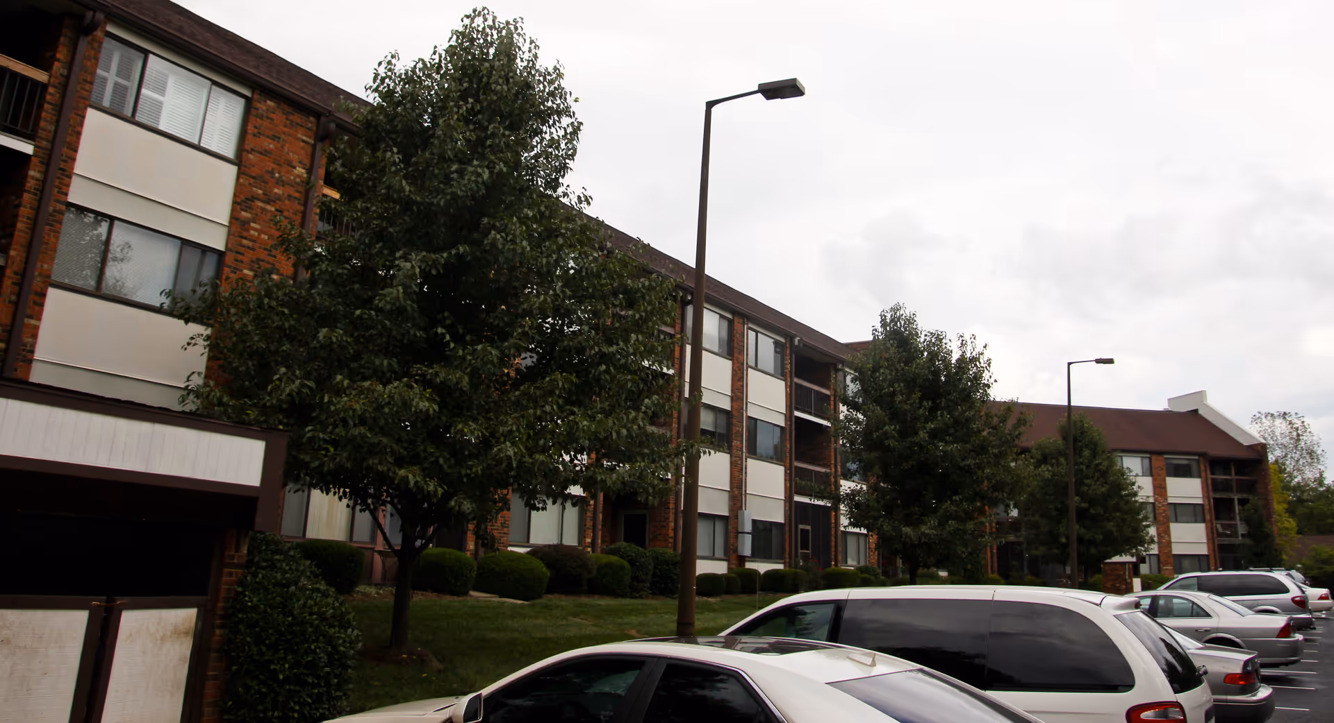 Exterior view of a multi-story senior living facility building with brick and white paneling, surrounded by trees and parked cars in the parking lot on a cloudy day.