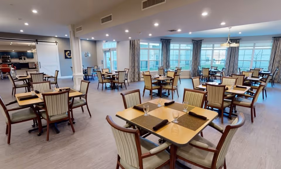 Spacious dining room with multiple square wooden tables, each set with four chairs that have striped upholstery. The tables are set with placemats, glasses, and rolled napkins. Large windows with patterned curtains allow natural light to fill the room. The room has recessed lighting and light-colored flooring. In the background, there is a kitchen area visible through a wide doorway.