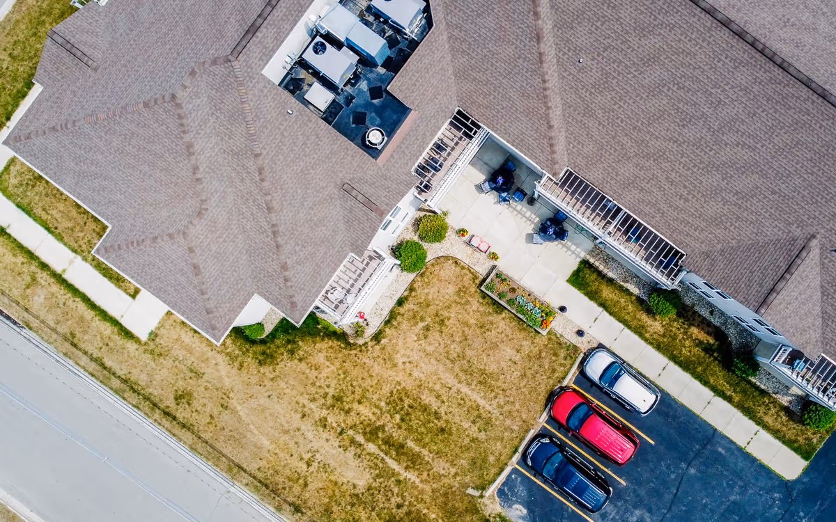 Aerial view of a senior living facility showing the roof of the building, a small parking lot with four cars, a sidewalk, and a grassy area with some small garden beds and patio furniture.