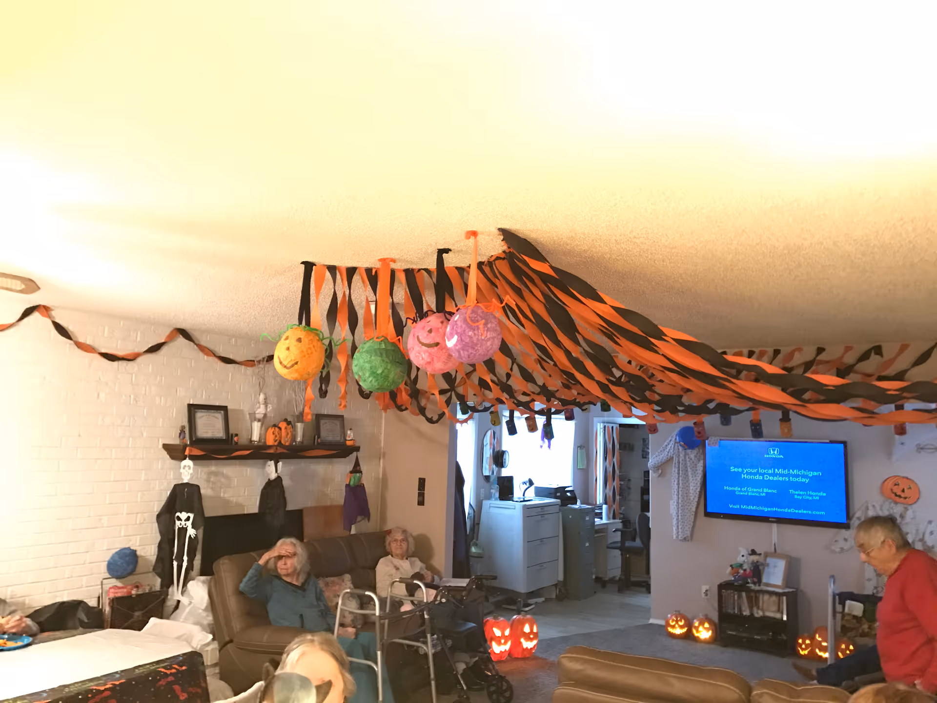 A living room decorated for Halloween with orange and black streamers hanging from the ceiling and various Halloween decorations including pumpkins and skeletons. Several elderly people are seated on couches and chairs, with one person using a walker. A television is mounted on the wall displaying a blue screen with text about local Honda dealers.