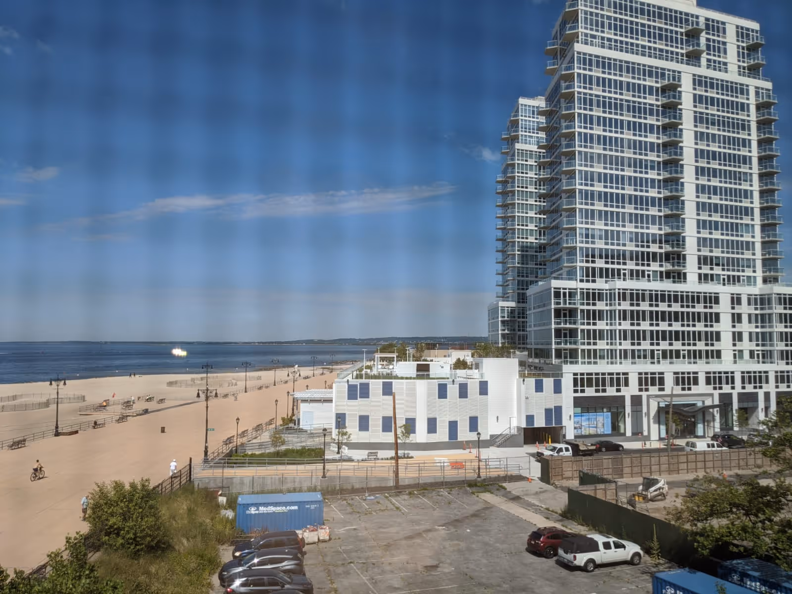 View of a beachfront area with a sandy beach, a boardwalk with benches and street lamps, and a large modern multi-story building with many windows near the shore. Several cars are parked in a lot in the foreground, and the sky is clear with a few clouds.