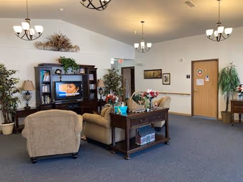 A spacious living room area in a senior living facility with beige upholstered armchairs and a sofa arranged around a dark wood entertainment center holding a television. The room features blue carpet, several potted plants, framed artwork on the walls, and multiple ceiling light fixtures. A wooden table with decorative items is placed behind the sofa.
