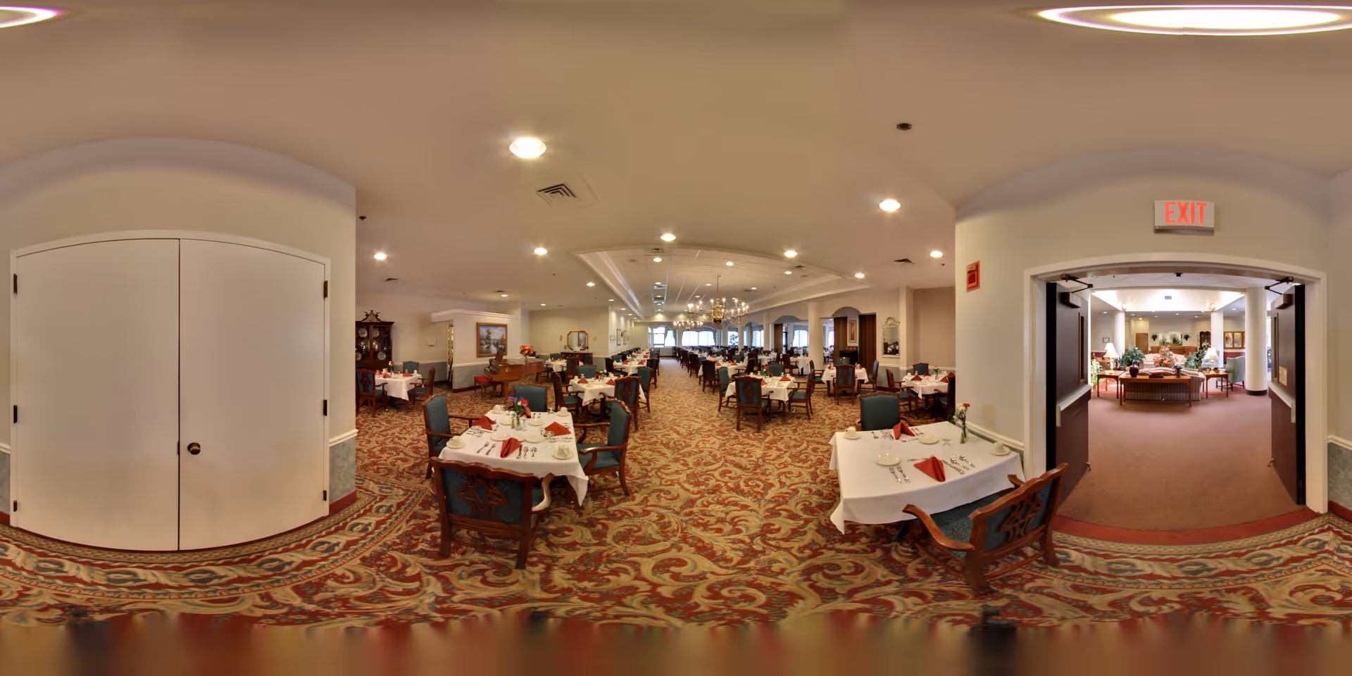 A spacious dining room in a senior living facility with multiple tables covered in white tablecloths, each set with red napkins, silverware, and small floral centerpieces. The room has patterned carpet flooring, chandeliers, and large windows letting in natural light. To the right, double doors lead to another room with more seating and plants.