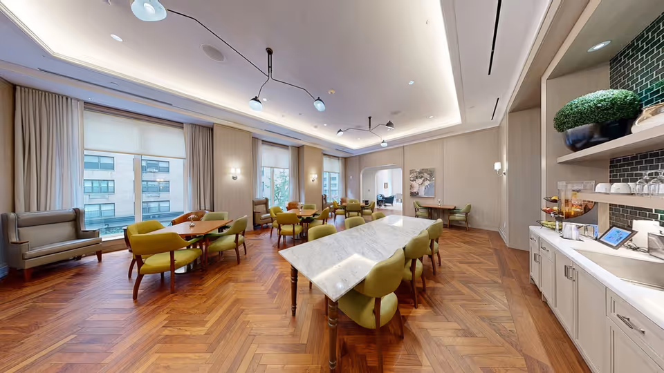A spacious, well-lit dining room with wooden herringbone flooring and large windows with beige curtains. The room features multiple tables with green upholstered chairs, a marble-topped table in the center, and a side counter with glassware, a beverage dispenser, and decorative plants. Modern ceiling lights and wall sconces provide additional lighting.