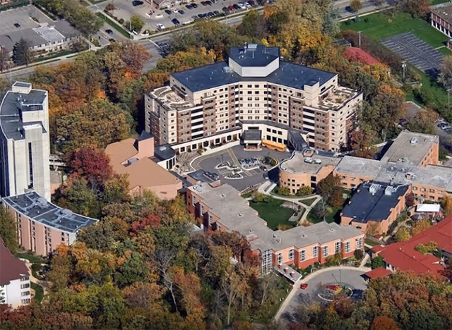 Aerial view of Oakwood Village University Woods senior living facility surrounded by trees with autumn foliage. The multi-story building complex includes a large central building with a curved facade, several smaller connected buildings, parking areas, and landscaped grounds.