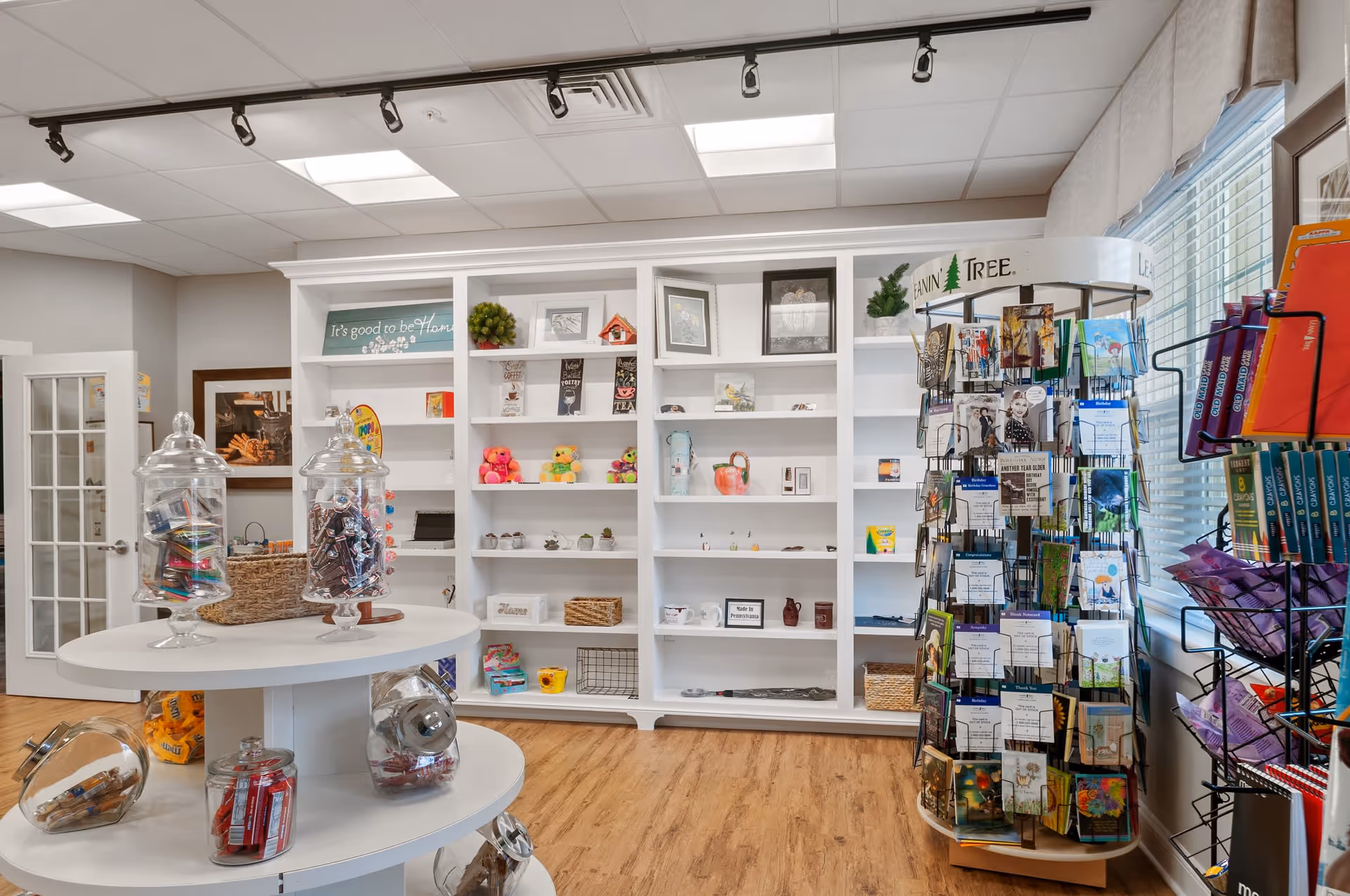 Interior view of a small gift shop area inside a senior living facility with white shelves displaying various decorative items, toys, and small gifts. A round display table holds jars filled with candy and snacks. A rotating card rack with greeting cards is positioned near a window with blinds. The floor is wooden, and the ceiling has recessed lighting.