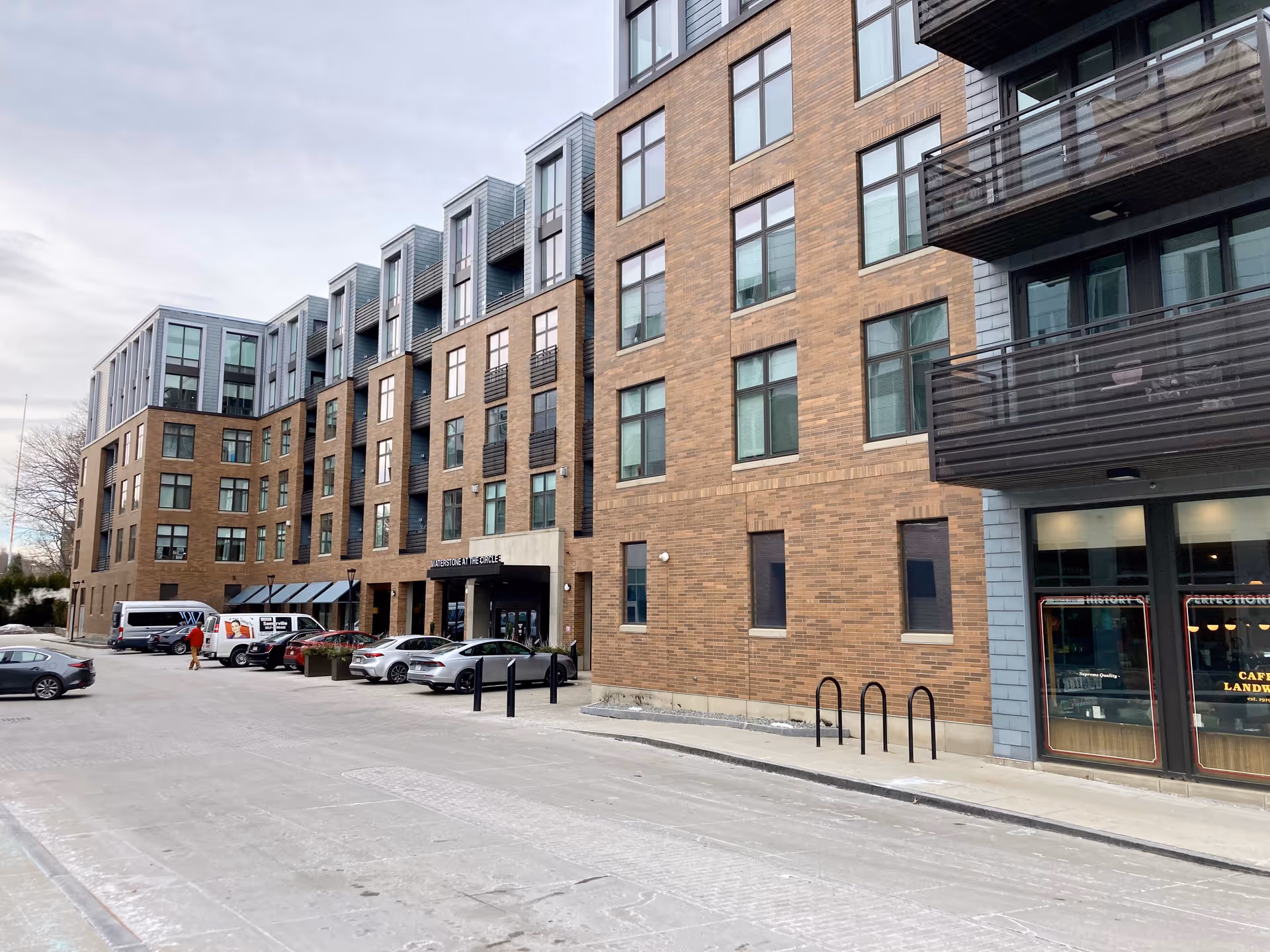 Exterior view of a modern multi-story brick building with large windows and balconies. Several cars are parked in front of the building near the entrance, which has a sign reading 'Waterstone at the Circle'. The sky is overcast.