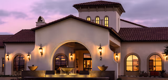 Exterior view of The Orchards Health Center building at dusk, featuring Spanish-style architecture with arched doorways and windows, warm lighting, and a tiled roof under a purple sky.