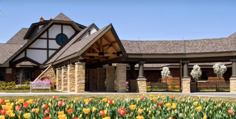 Exterior view of Mcgregor Assisted Living facility featuring a covered entrance with stone pillars, a peaked roof, and a garden with blooming red and yellow tulips in the foreground under a clear blue sky.