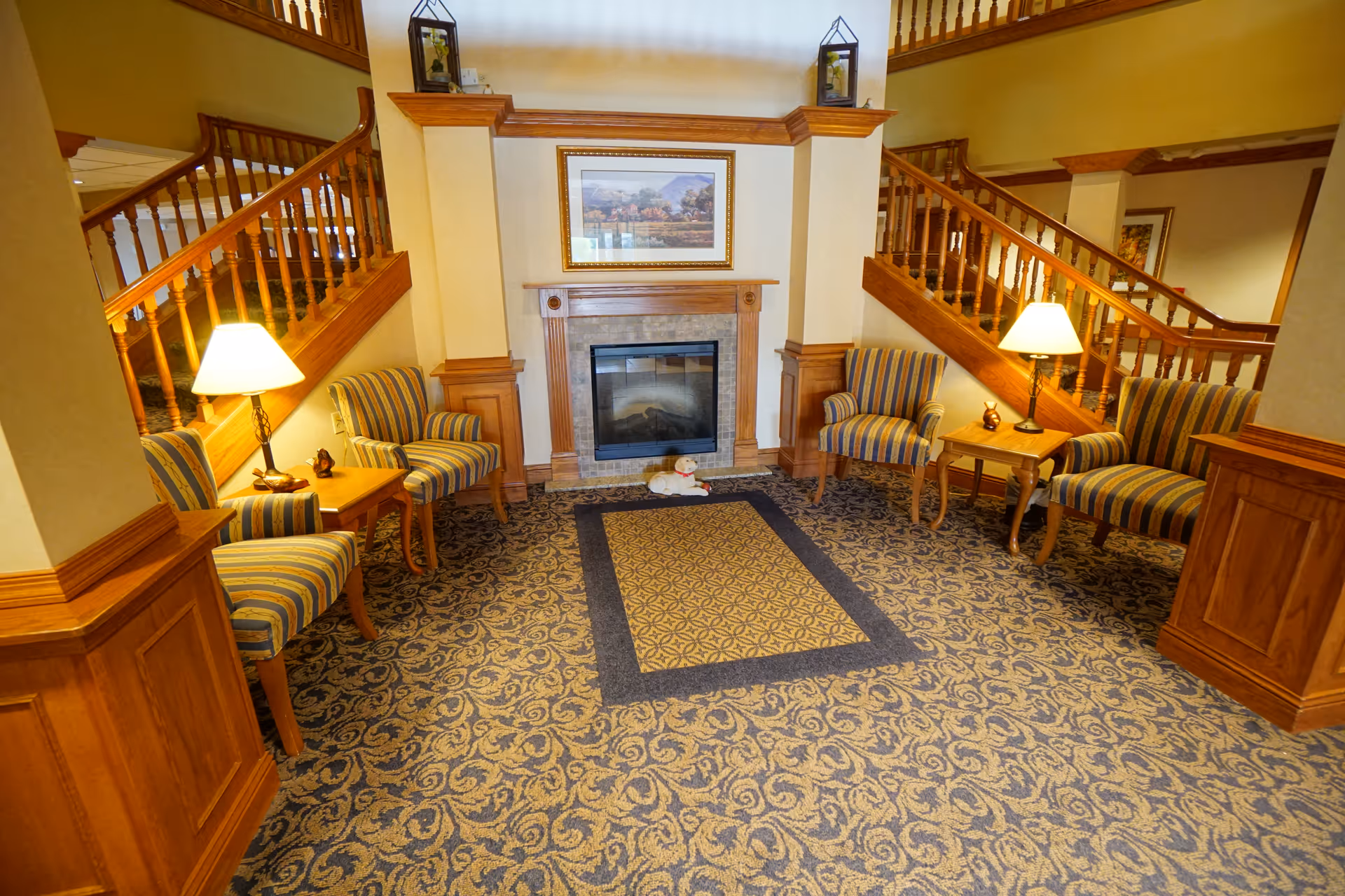 A cozy seating area in a senior living facility with a fireplace in the center, flanked by two wooden staircases. The room features patterned carpet, two striped armchairs on each side, two small wooden tables with lamps, and a framed landscape painting above the fireplace. A small stuffed dog toy is placed on the carpet in front of the fireplace.
