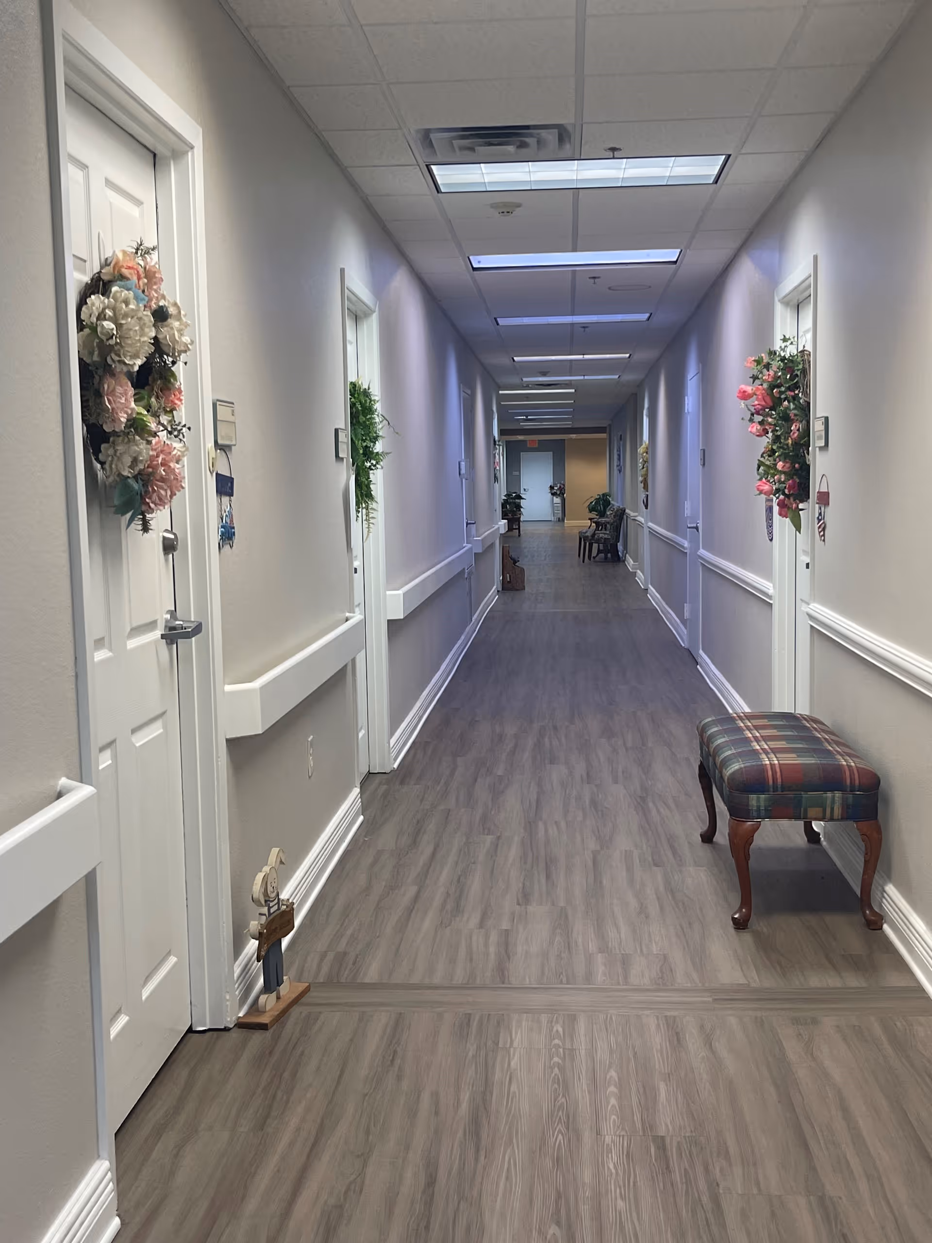 A long, well-lit hallway in a senior living facility with light-colored walls and wood-patterned flooring. Several doors line both sides of the hallway, each decorated with floral wreaths. A plaid upholstered bench is positioned against the right wall, and a small decorative figure stands near a door on the left. The ceiling has recessed lighting panels.