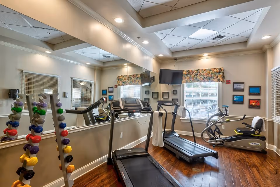 A small fitness room with wooden floors, featuring two treadmills, a recumbent exercise bike, and a rack of colorful dumbbells. The room has large windows with floral valances, a wall-mounted TV, and several framed pictures on the walls. A large mirror covers one wall, reflecting the equipment and windows.
