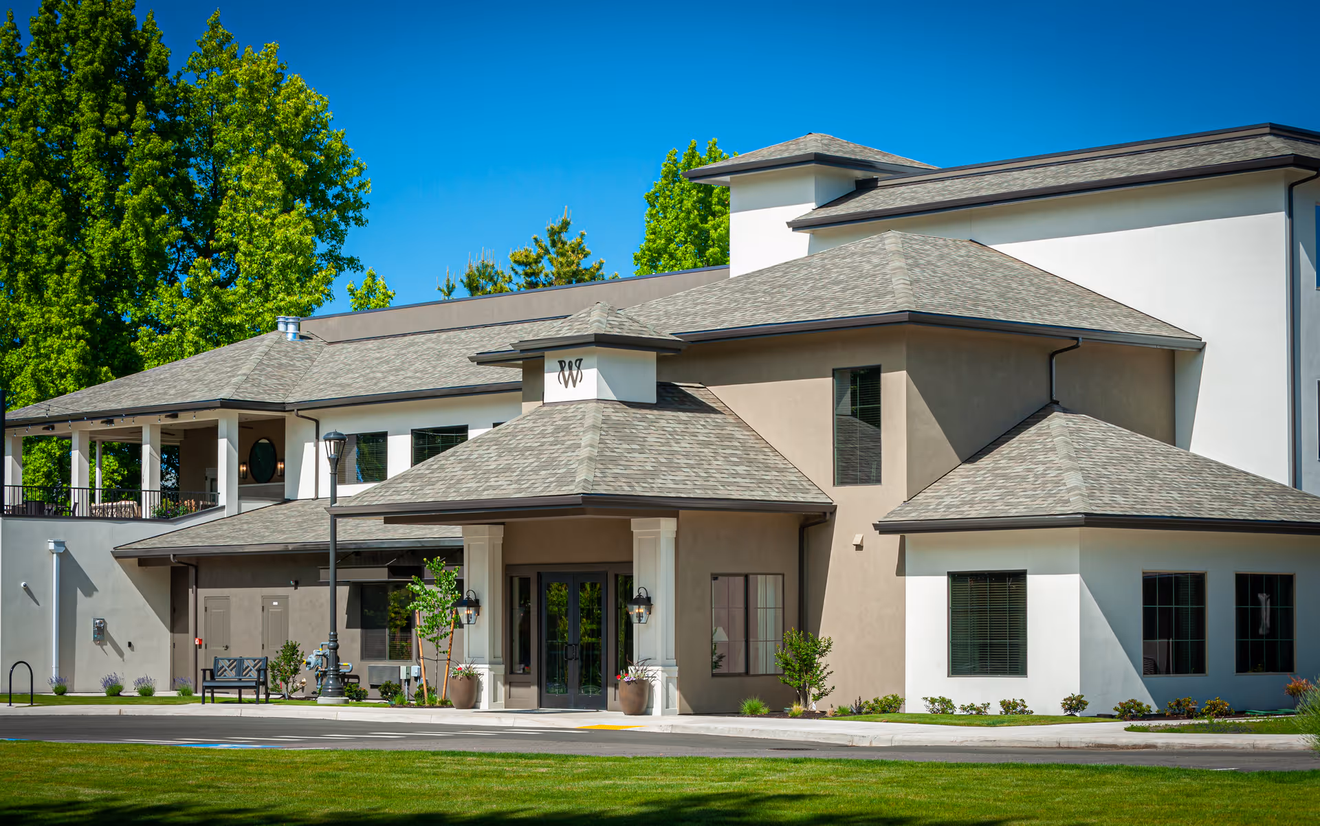 Exterior view of a modern senior living facility building with beige and white walls, multiple roof levels, large windows, and a covered entrance. The building is surrounded by green grass and trees under a clear blue sky.