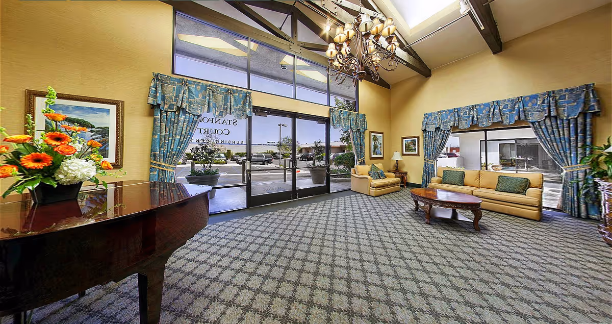 A spacious and well-lit common area with large windows and glass doors leading outside. The room features a patterned carpet, two beige sofas with green cushions, a wooden coffee table, a grand piano with a floral arrangement on top, framed artwork on the walls, and blue patterned curtains. A chandelier hangs from the ceiling with exposed wooden beams.