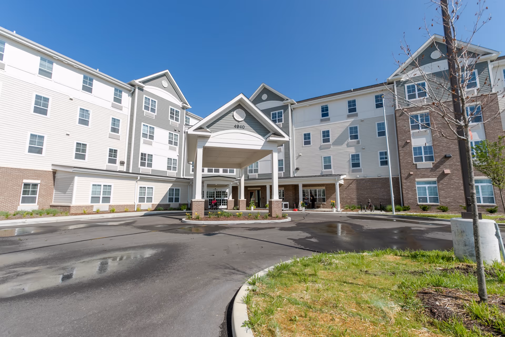 Exterior view of a multi-story senior living facility building named Oasis at 56th with a covered entrance, multiple windows, and a driveway in front. The sky is clear and blue, and there is some landscaping with grass and small trees around the building.