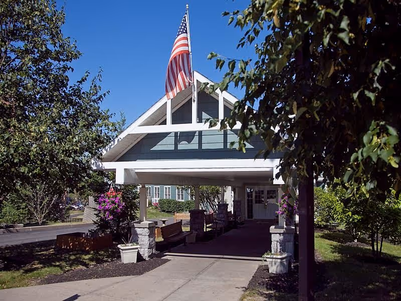 Entrance of a senior living facility with a covered walkway supported by stone pillars, an American flag flying on top, benches along the walkway, and surrounding trees and plants under a clear blue sky.