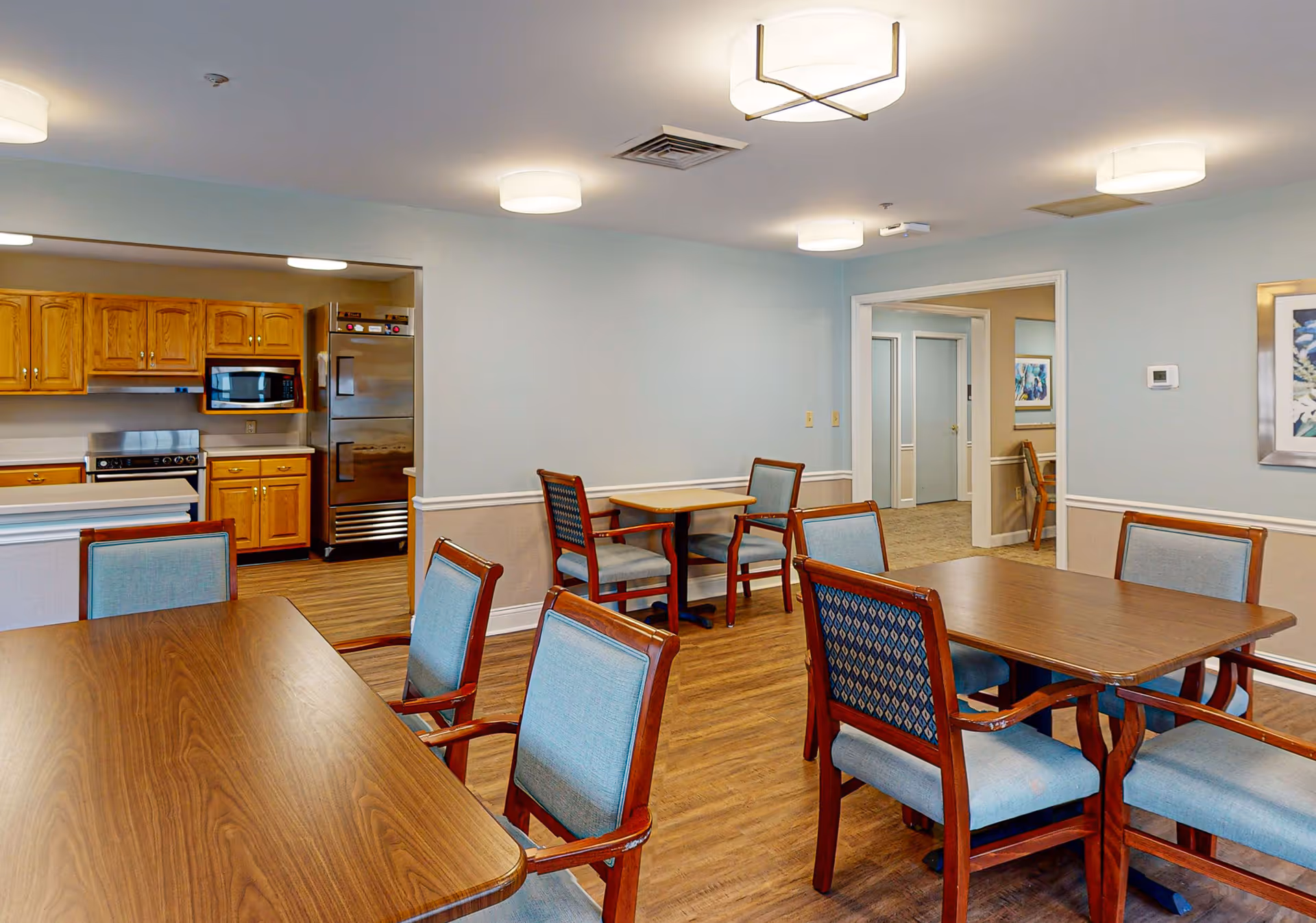 A dining area in a senior living facility with wooden tables and chairs upholstered in light blue fabric. The room has light blue walls with white trim and wood flooring. In the background, there is a kitchen area with wooden cabinets, a microwave, a stove, and a stainless steel refrigerator. Ceiling lights provide illumination.