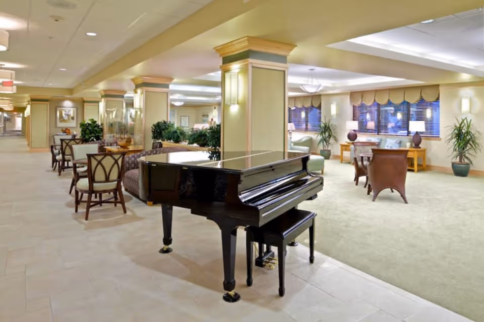Spacious senior living facility common area with a black grand piano in the foreground, surrounded by comfortable seating including chairs and sofas. The room features beige walls with decorative columns, large windows with blinds, potted plants, and soft lighting fixtures creating a warm and inviting atmosphere.
