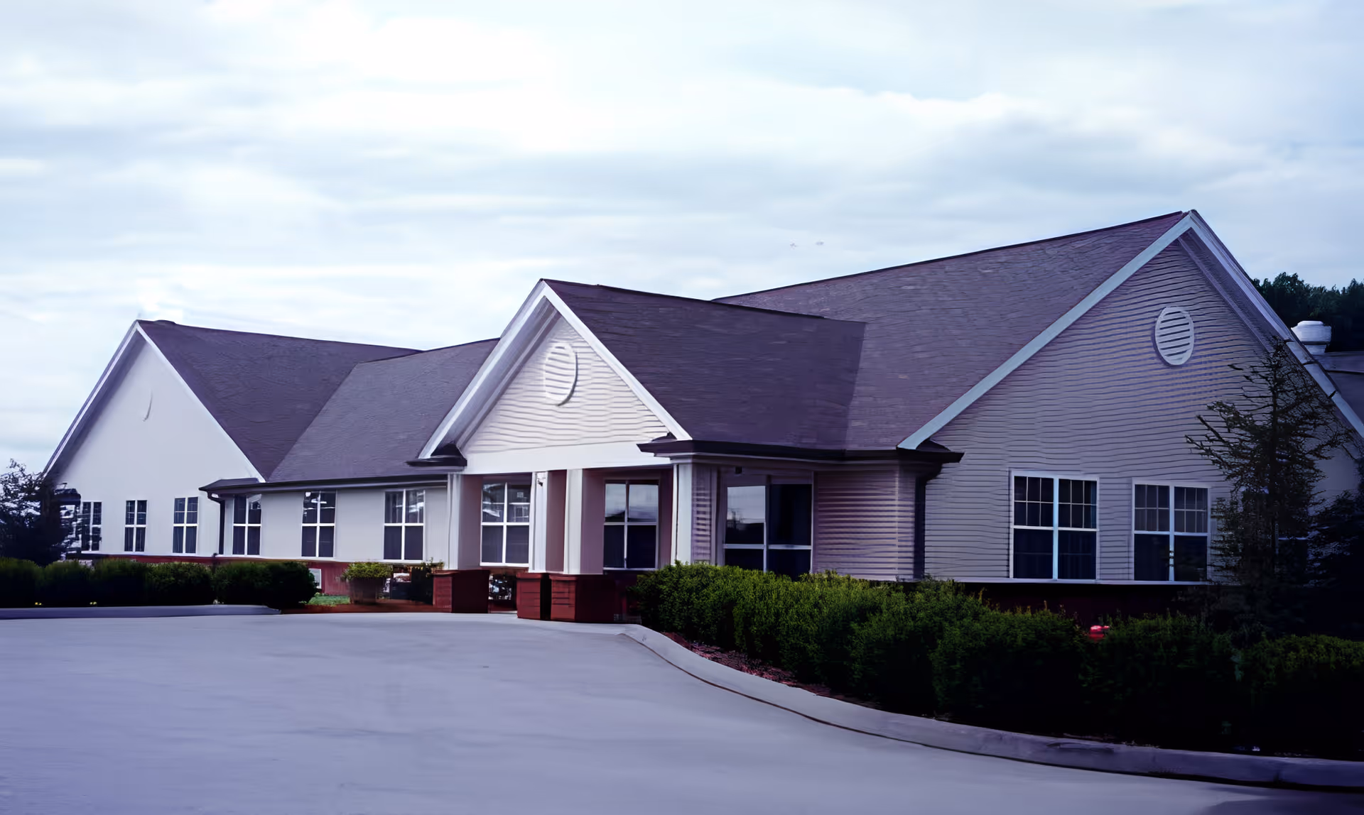 Exterior view of a single-story building with a pitched roof, multiple windows, and a covered entrance. The building is surrounded by neatly trimmed bushes and a paved driveway leading up to the entrance. The sky is partly cloudy.