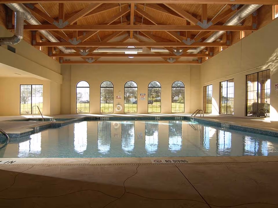 Indoor swimming pool with clear water, surrounded by beige walls and large arched windows letting in natural light. The ceiling features exposed wooden beams and metal ductwork. There are 'No Diving' signs visible on the pool edge and walls.