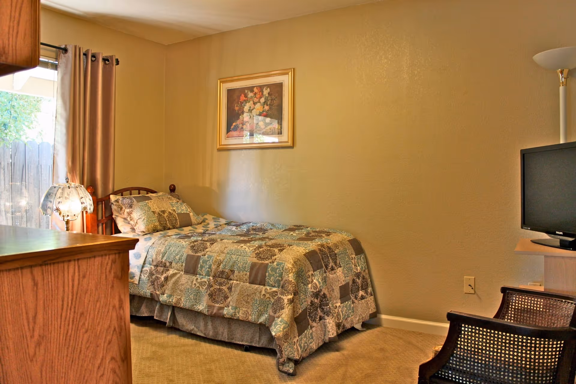A cozy bedroom with a single bed covered in a patterned quilt. There is a wooden dresser on the left, a window with beige curtains, a framed floral painting on the wall above the bed, a floor lamp, a TV on a stand, and a wooden chair with a woven backrest.