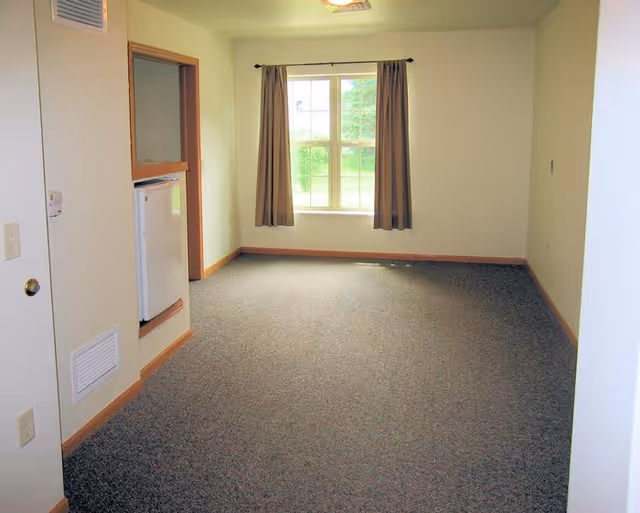 Empty room with beige walls, a window with brown curtains, gray carpeted floor, and a small built-in white mini fridge on the left wall.
