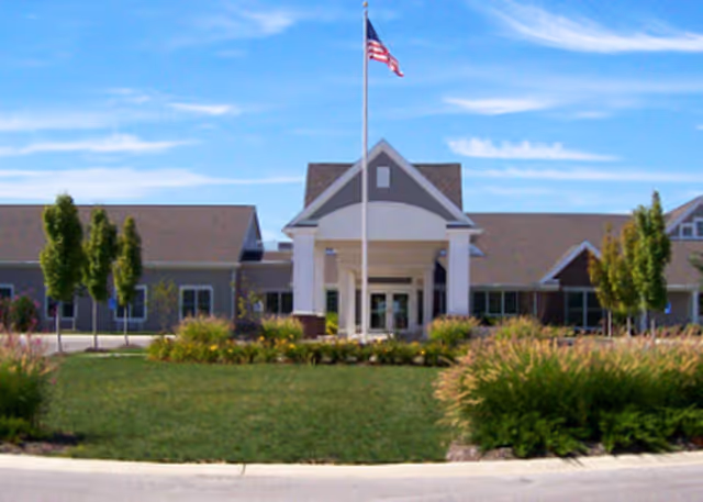 Front exterior view of Briarwood Village facility with a central entrance featuring white columns and a peaked roof. An American flag is flying on a flagpole in front of the building, surrounded by green lawn and landscaping with bushes and small trees under a blue sky with some clouds.