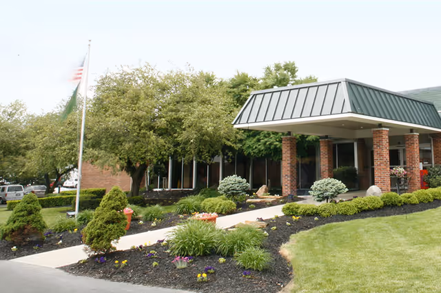 Exterior view of Monterey Care Center showing the entrance with a covered drop-off area supported by brick columns, surrounded by well-maintained landscaping including bushes, small trees, flowers, and a neatly trimmed lawn. An American flag is visible on a flagpole to the left.