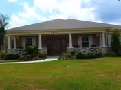 Single-story building with a covered front porch, surrounded by green grass and shrubs under a partly cloudy sky.