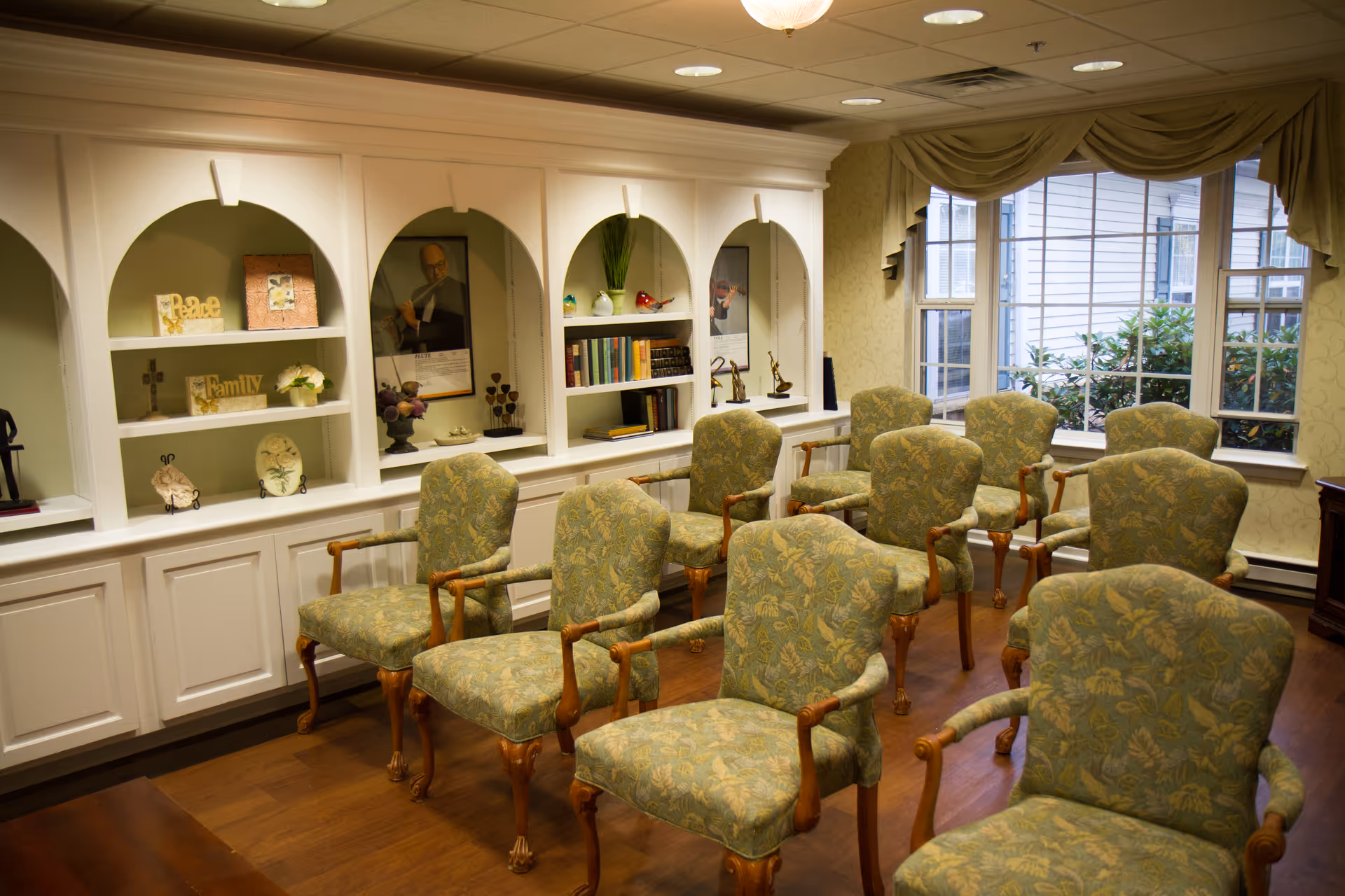 A cozy room with multiple upholstered armchairs arranged in rows facing a built-in white shelving unit with decorative items, books, and framed pictures. A large window with draped curtains allows natural light to enter, and the room has wooden flooring.