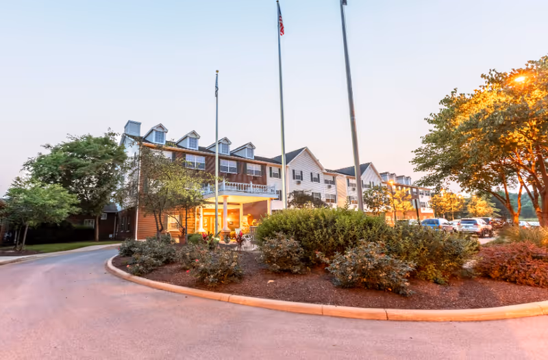 Exterior view of a multi-story senior living facility building with a driveway, landscaped bushes, trees, and flagpoles in front during early evening light.