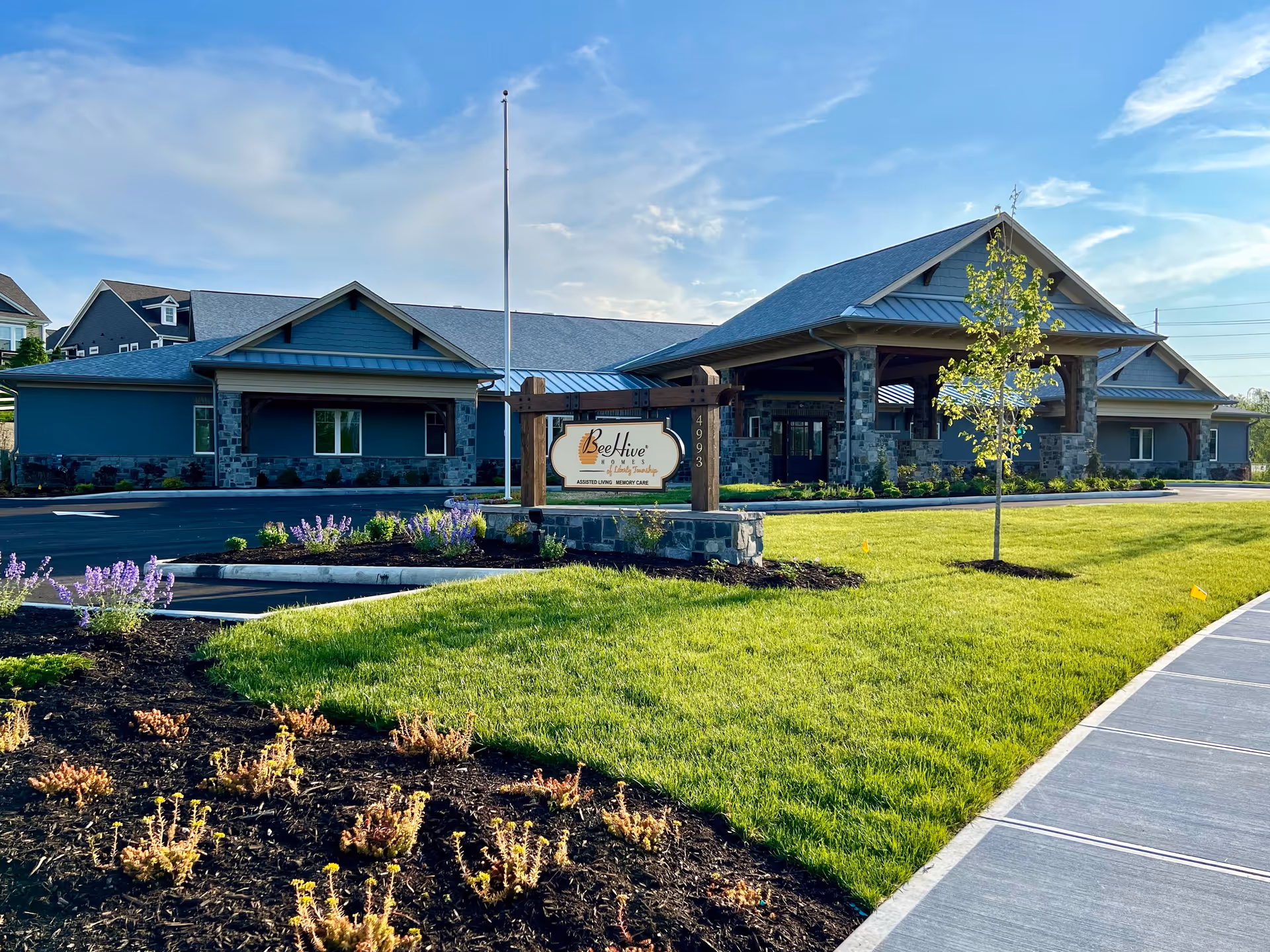 Exterior view of Beehive Homes of Liberty Township building with a landscaped lawn, flower beds, a young tree, and a clear blue sky in the background. The building features stone and wood accents with a covered entrance and a sign displaying the facility's name.