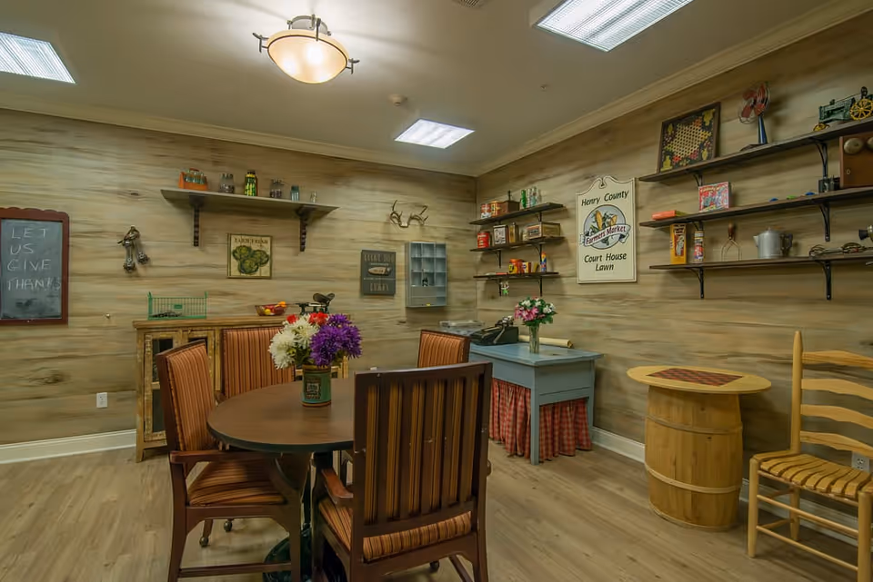 A cozy room with wooden walls and flooring featuring a round table with four striped cushioned chairs and a vase of colorful flowers in the center. The room has shelves on the walls holding various decorative items and vintage signs, including one that reads 'Henry County Farmers Market Court House Lawn.' There is a small blue table with a typewriter and a wooden barrel with a checkerboard on top, along with a wooden chair. The lighting includes a ceiling light fixture and fluorescent panels.